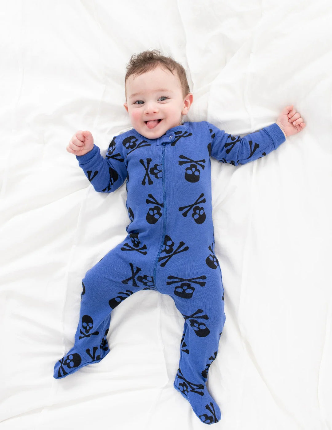 A baby lies on a white bed, wearing Leveret Halloween Pajamas with a black skull and crossbones pattern. The best-selling blue onesie has the baby smiling with their arms and legs outstretched.